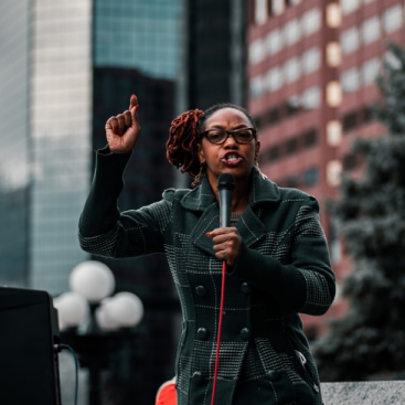 A black woman wearing a grey striped coat speaking passionately with a microphone.